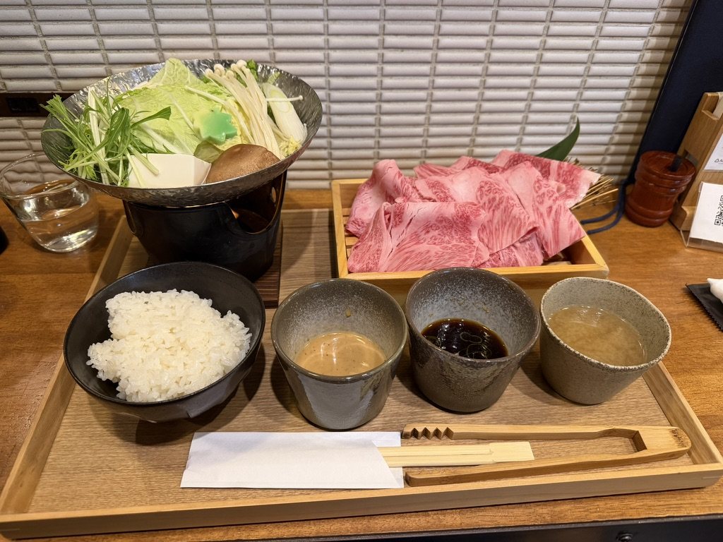 A tray of wagyu slices, vegetables in a hot pot, rice, and dipping sauces