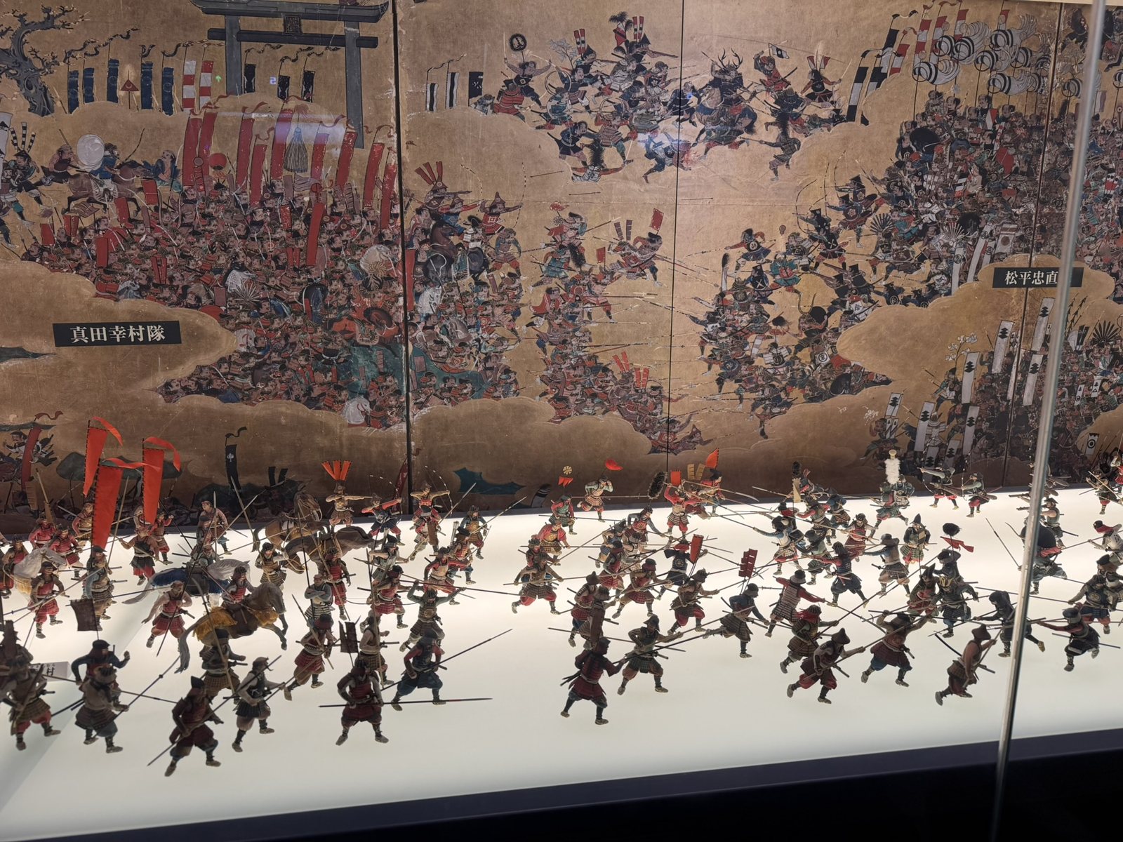A diorama of samurai fighting in front of a painted folding screen of the Siege of Osaka
