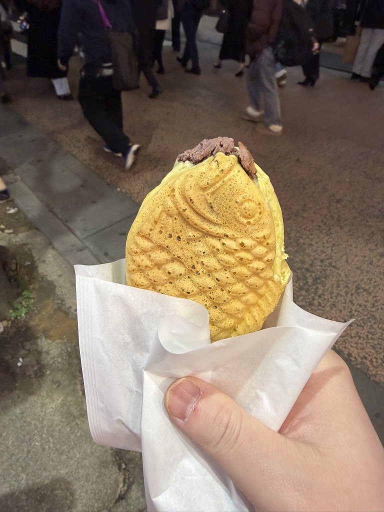 A fish-shaped taiyaki filled with sweet bean paste, held over a busy street