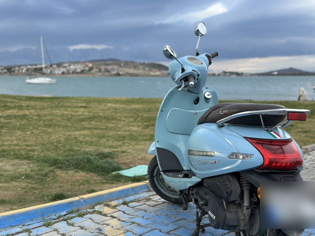 A pale blue scooter parked by the sea at Ayvalık, with a sailboat and the far shore behind